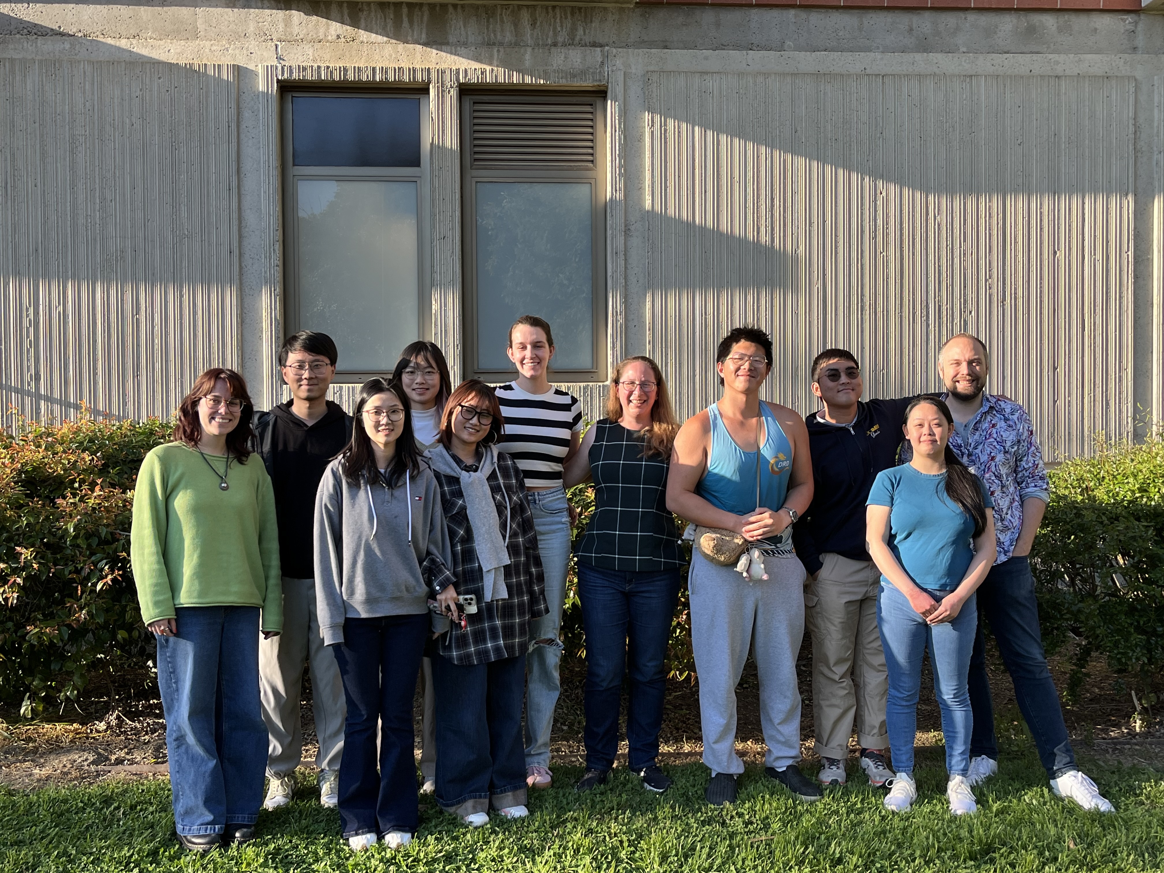 Outdoor group photo of 12 people standing on grass in front of a beige building